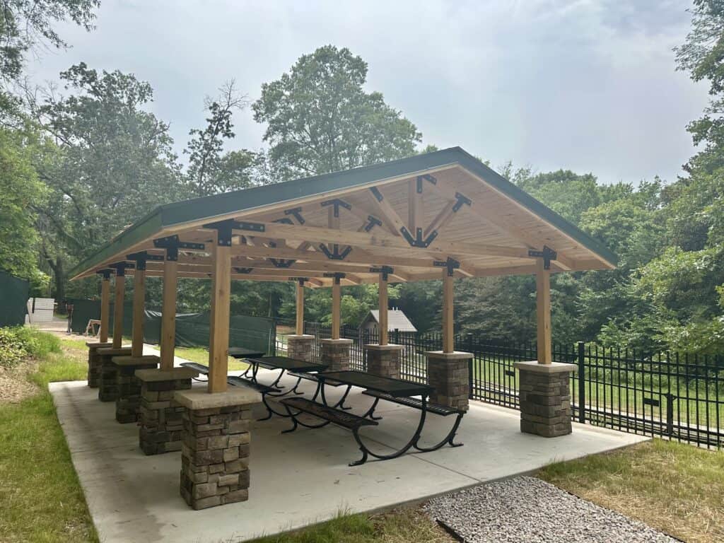 Modern park pavilion shelter with metal roof, concrete pad, and picnic tables in a public park setting