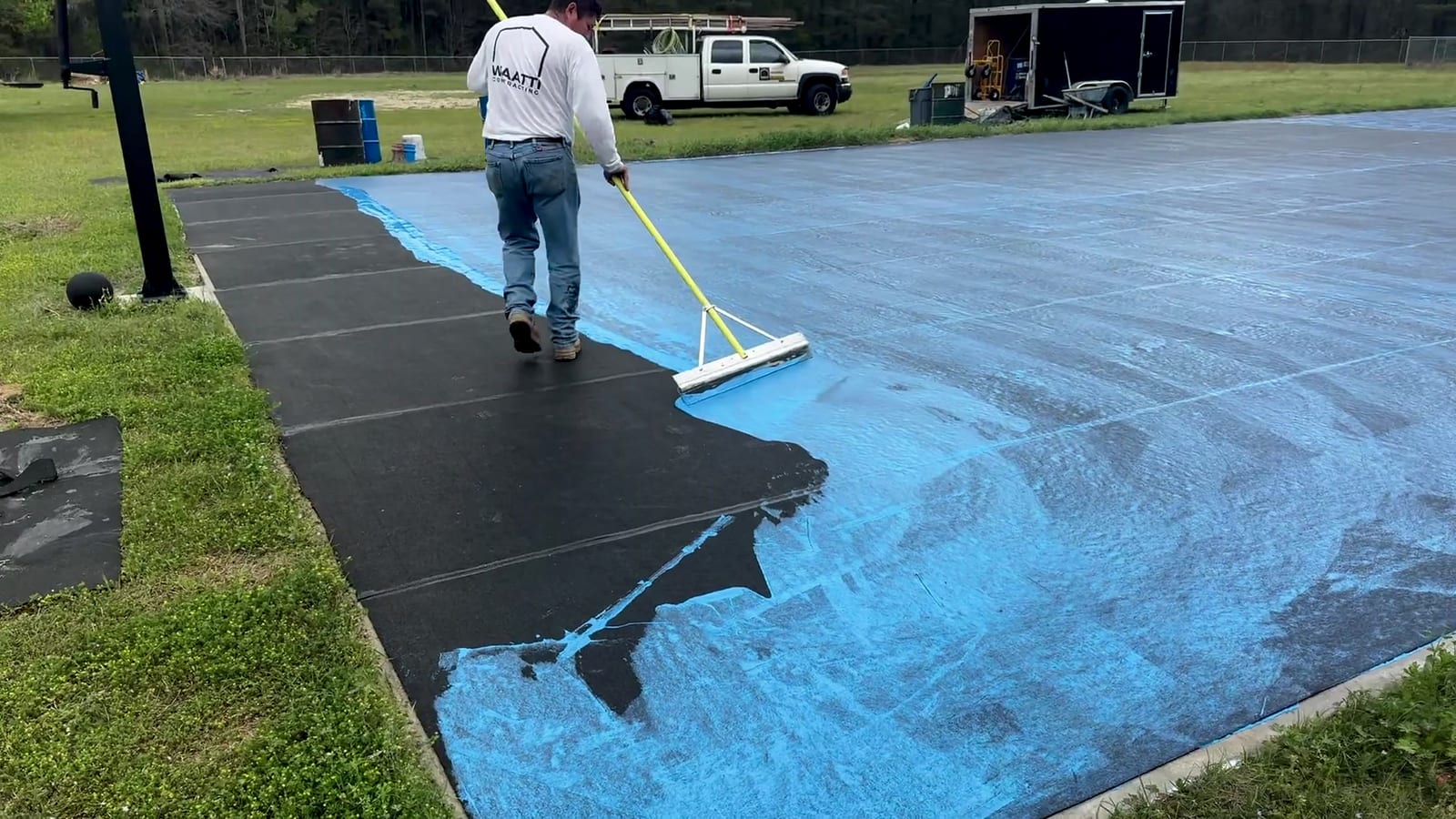 Crew member in a Waatti long-sleeve shirt squeegeeing blue acrylic over black resurfacer on a new asphalt basketball court; work truck beyond — East Arcadia, NC