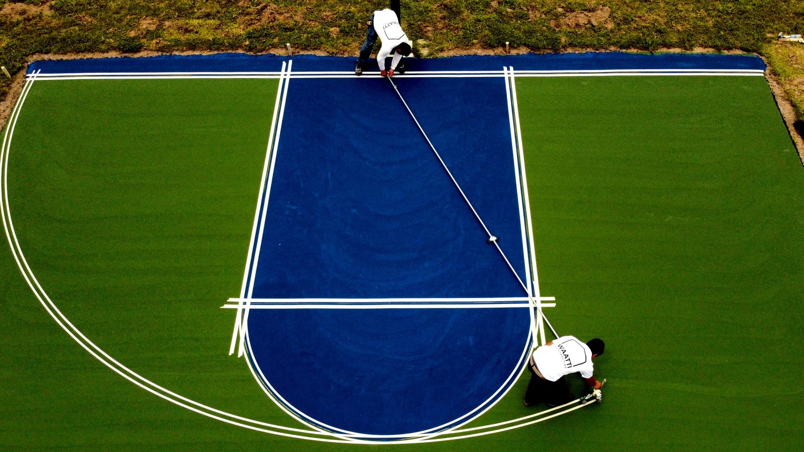 Drone view of two crew members taping and painting white key lines on a new blue and green basketball court — East Arcadia, NC