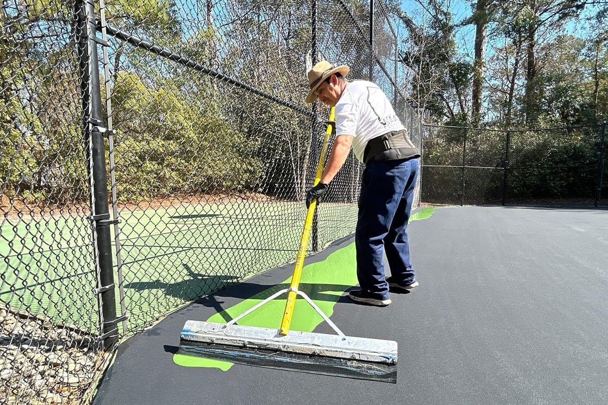 Waatti crew member pulling green acrylic color coat across a tennis court with a squeegee, fence and pines behind — Wilmington, NC
